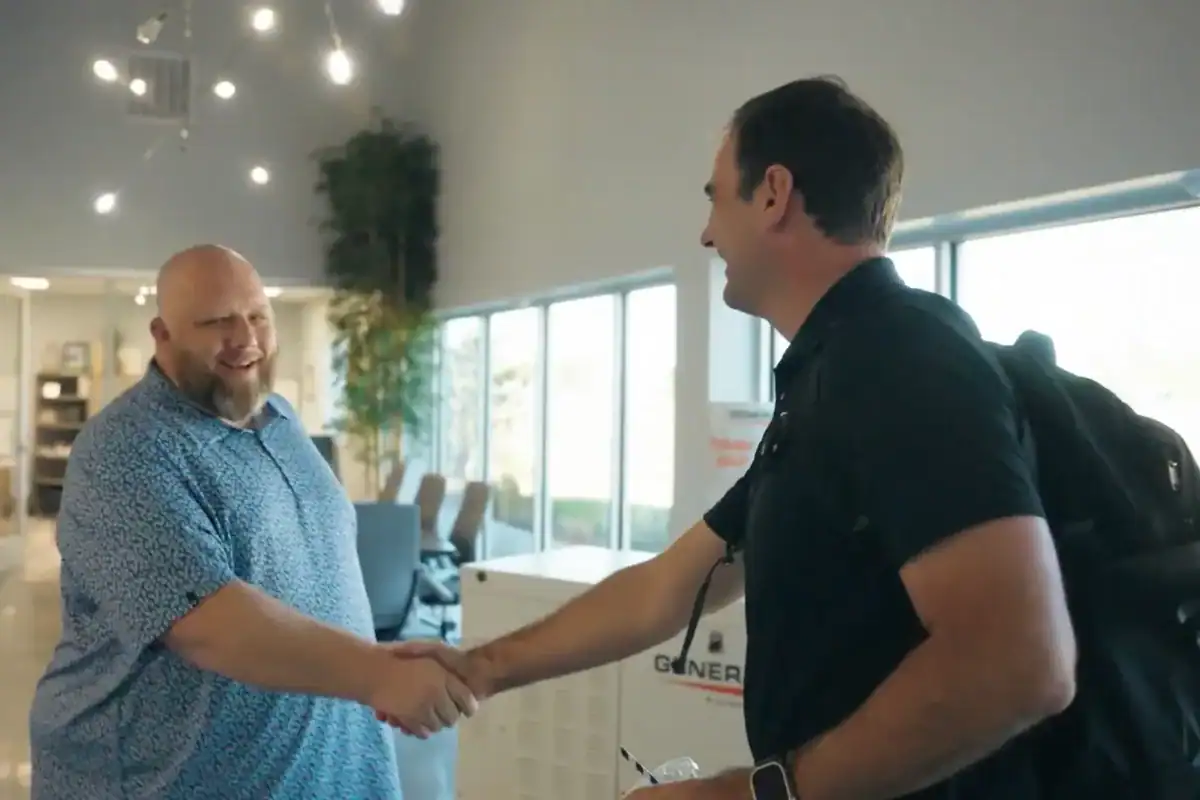 A Grasten team member and a Canter team member shake hands in an office lobby, a Generac standby generator on display behind them